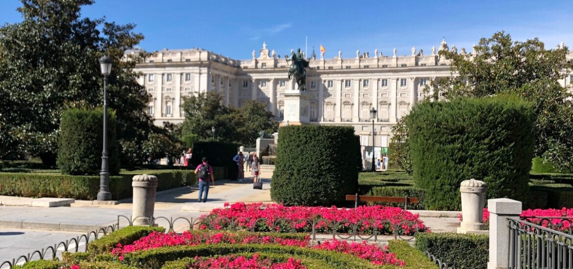 Small garden of short green hedges and hot pink flowers with a palace in the background on a clear day.