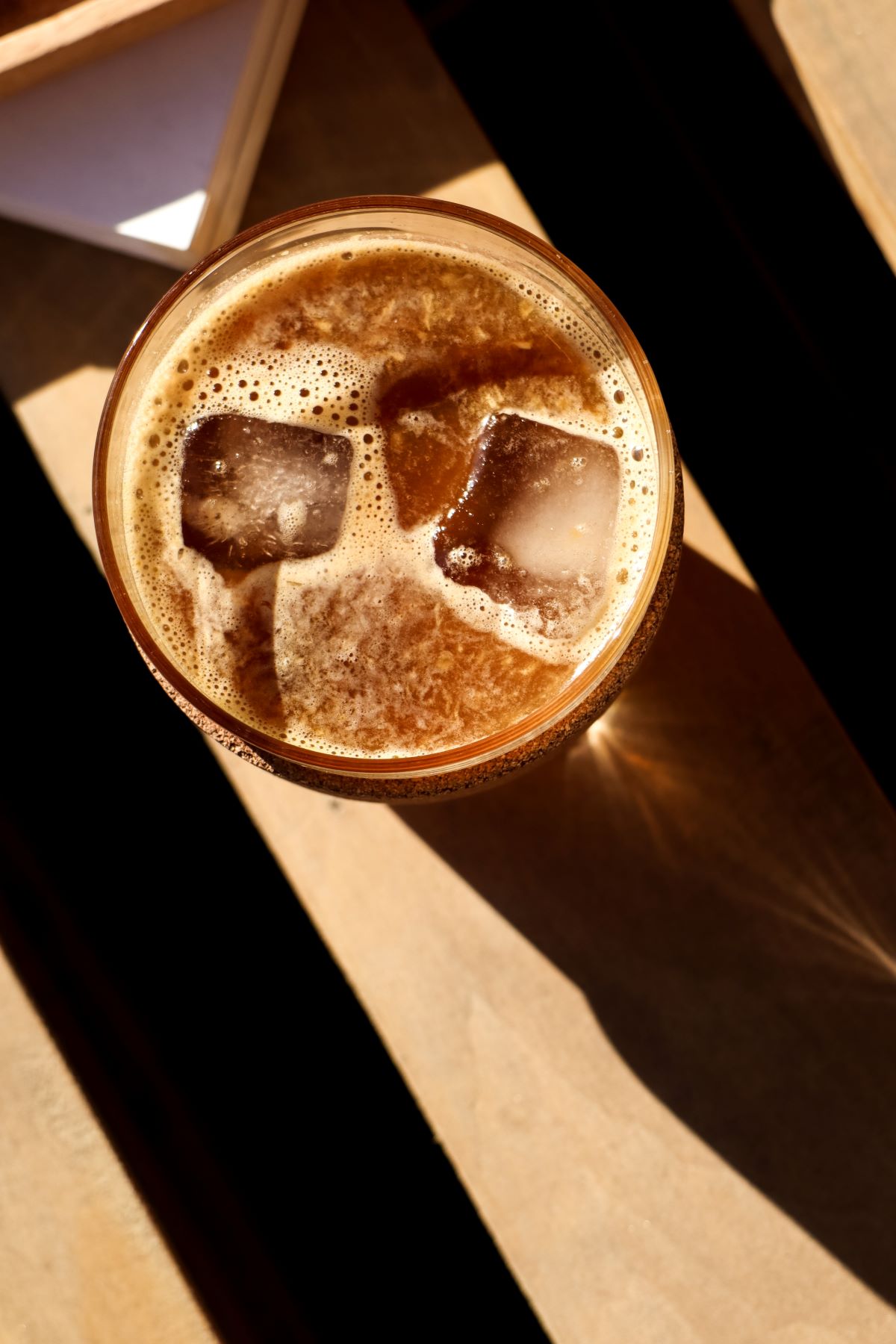 A drink with ice cubs sitting on a table with the sun reflecting on it.