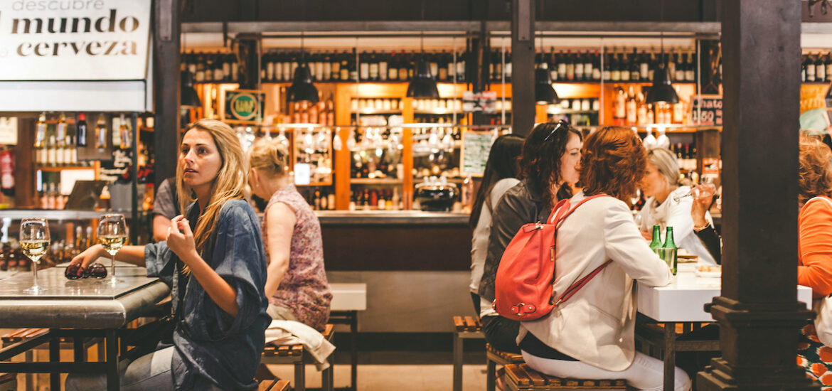 People seated at high tables inside a food hall eating tapas and drinking wine