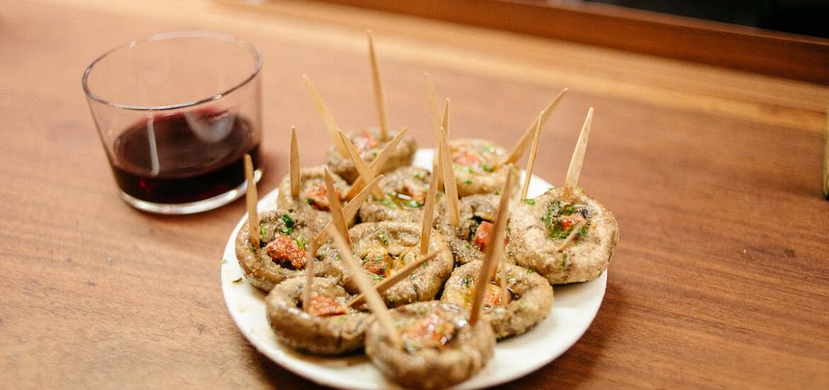 Plate of grilled mushrooms with chorizo next to a short glass of red wine.