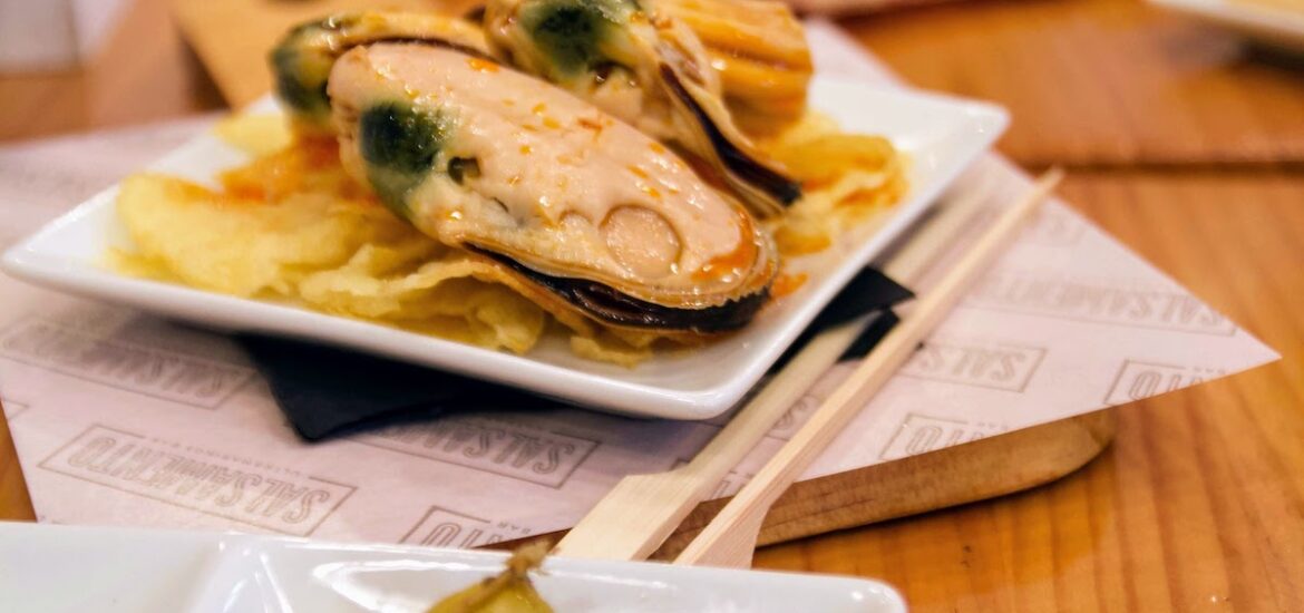 Two small square dishes of mussels and green olives on a wooden surface