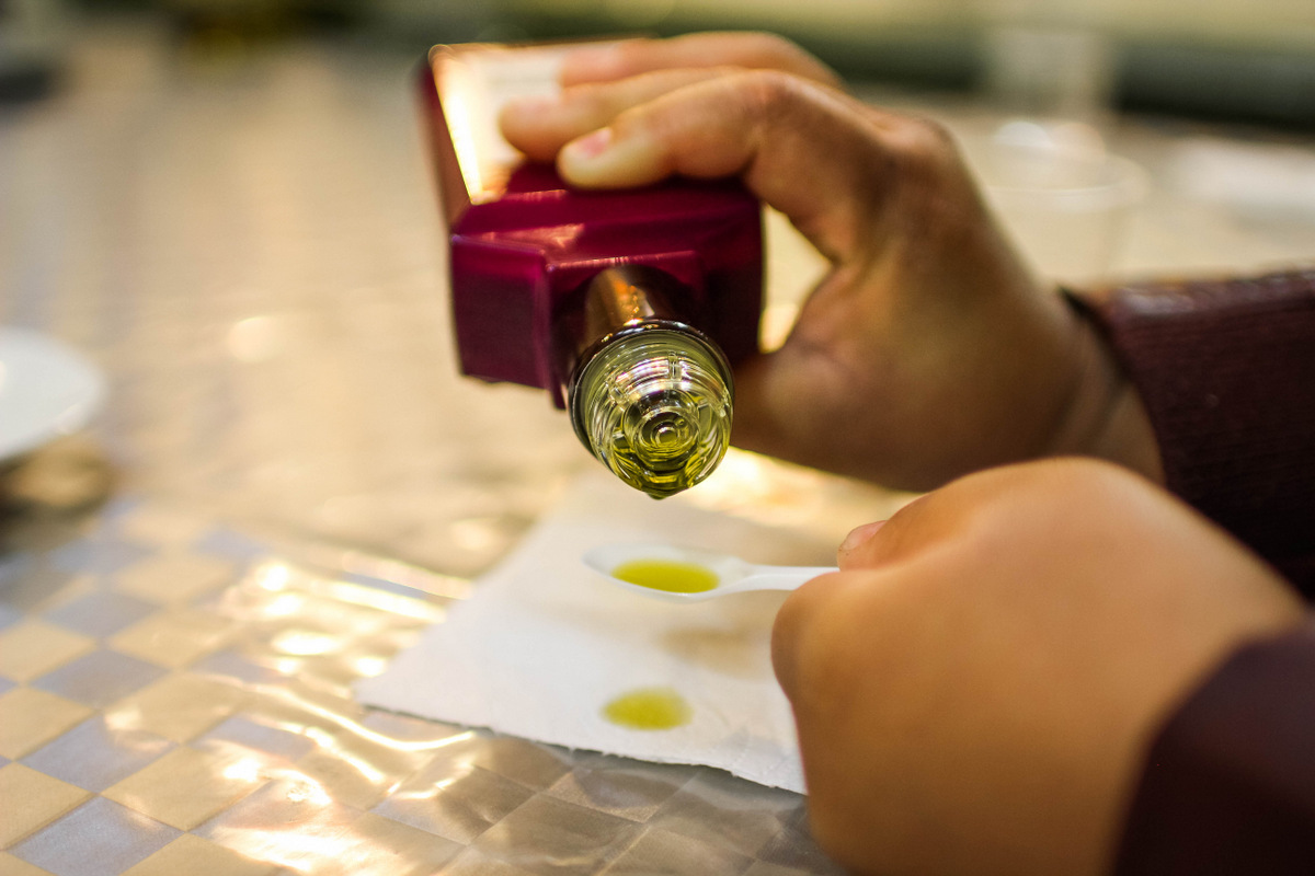 Close up of a person's hands pouring olive oil from a glass bottle onto a small white spoon.