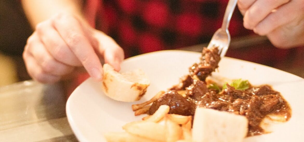 Close up of a person in a red shirt eating a meat dish while using a piece of bread to soak up the sauce.