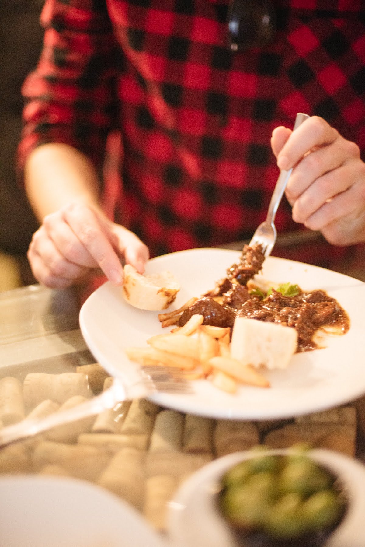 Person in a red and black checkered shirt eating a meat dish with a small piece of bread