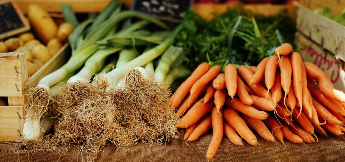 A table at a farmers market filled with leeks and carrots.