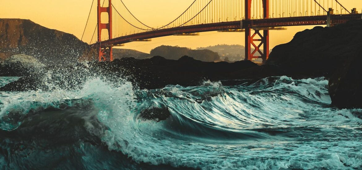 View of the Golden Gate Bridge in San Francisco from a beach. The waves are crashing and the sky is golden yellow.