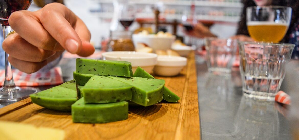 Person reaches to take a bite of green cheese at a cheese store with beer on the table next to them