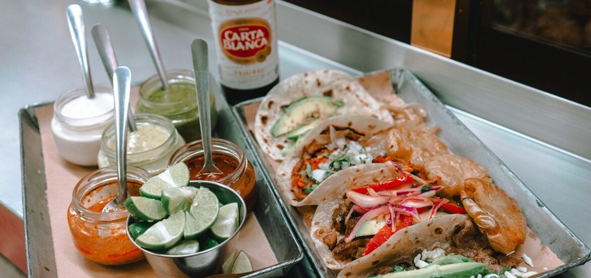 A tray of street tacos with limes, sauces, and beer