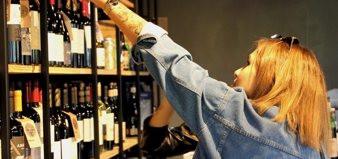 A woman in a jean shirt points to a tall shelf of wine for sale