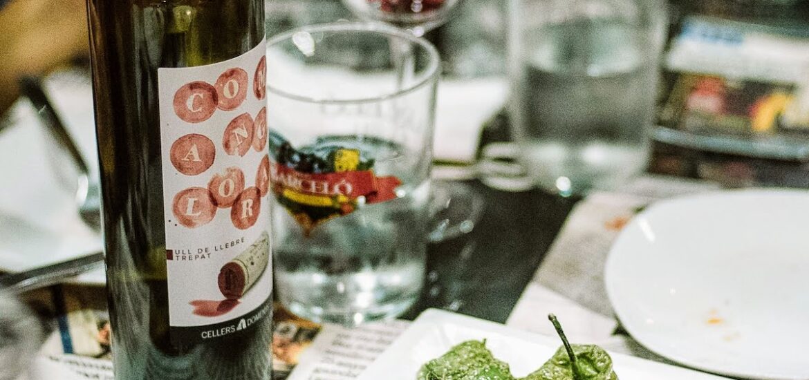 Small green peppers on a white plate beside a bottle of wine on a crowded tabletop