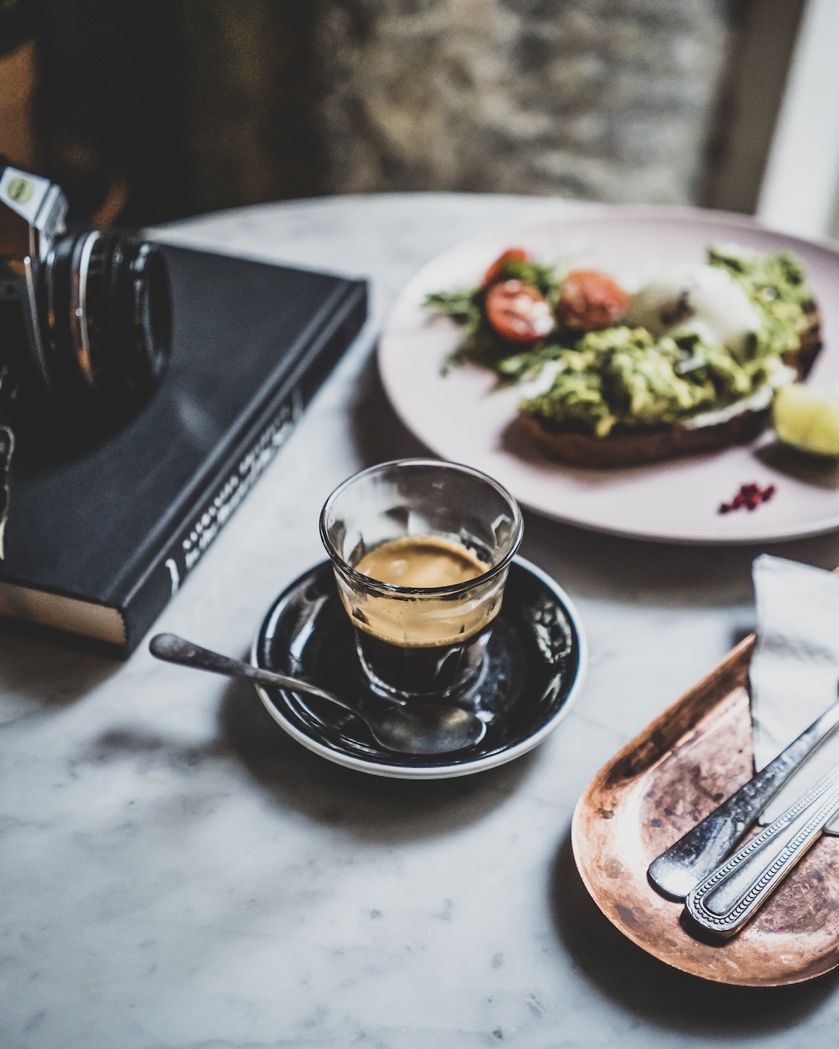 Clear glass of espresso next to a book and a plate of avocado toast on a white tabletop.