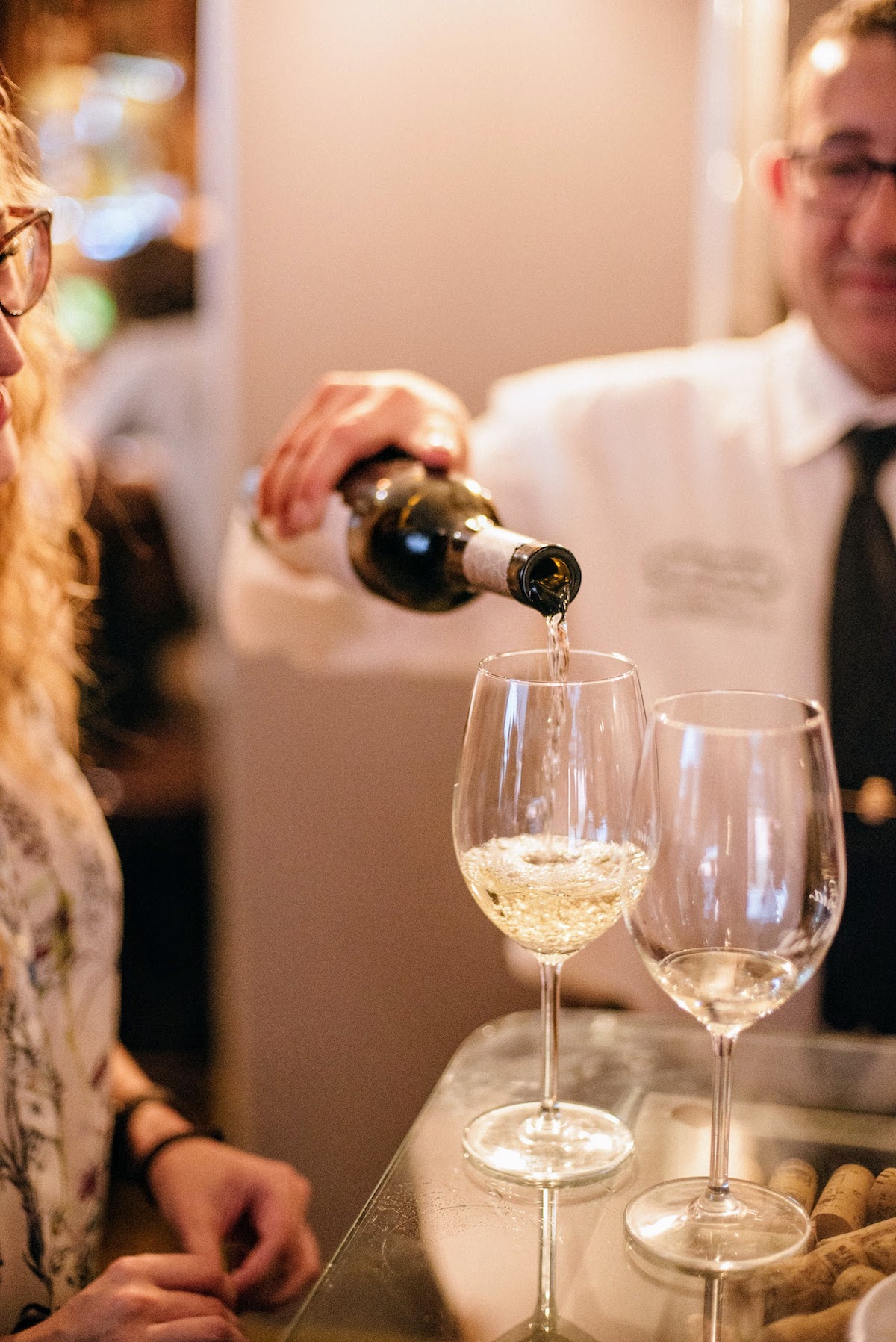 A bartender in a white shirt and tie pouring two glasses of white wine