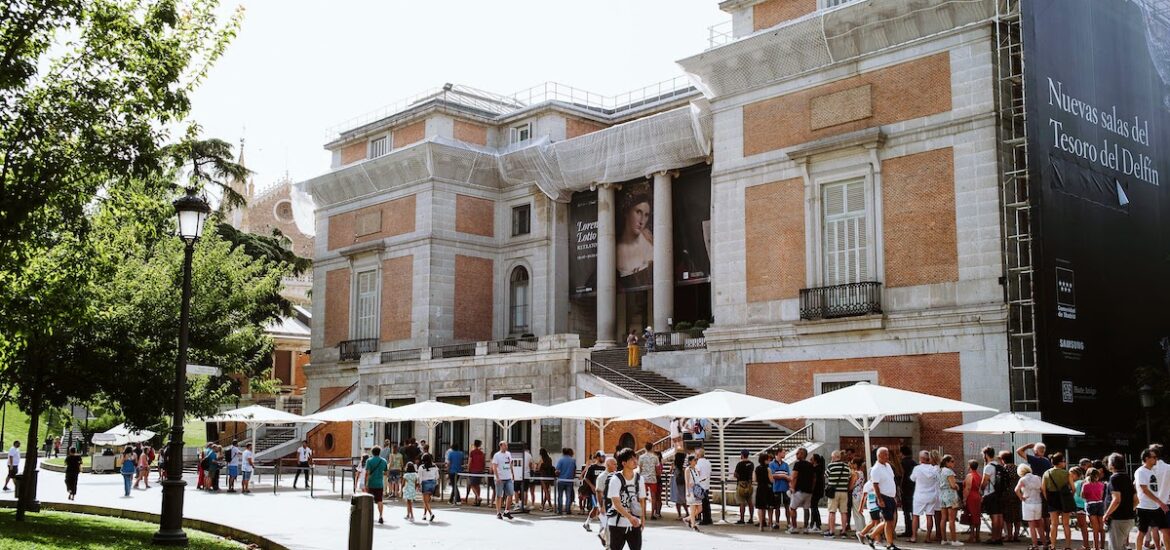 Exterior of a large brick and stone building with people standing in line under shady umbrellas outside.