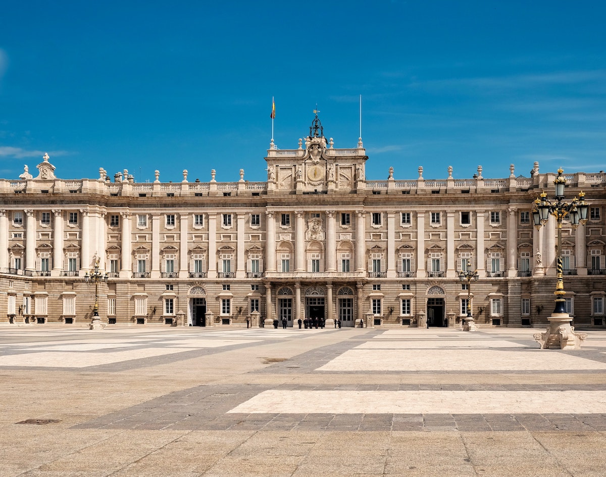 Exterior of a large, grandiose royal palace as seen from across an expansive plaza on a clear day.