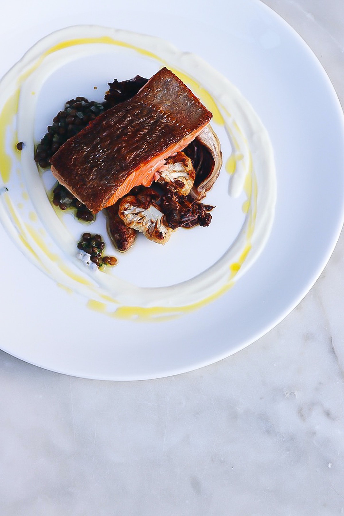 Overhead shot of a blackened salmon fillet and cauliflower on a white plate artfully garnished with a white and yellow sauce