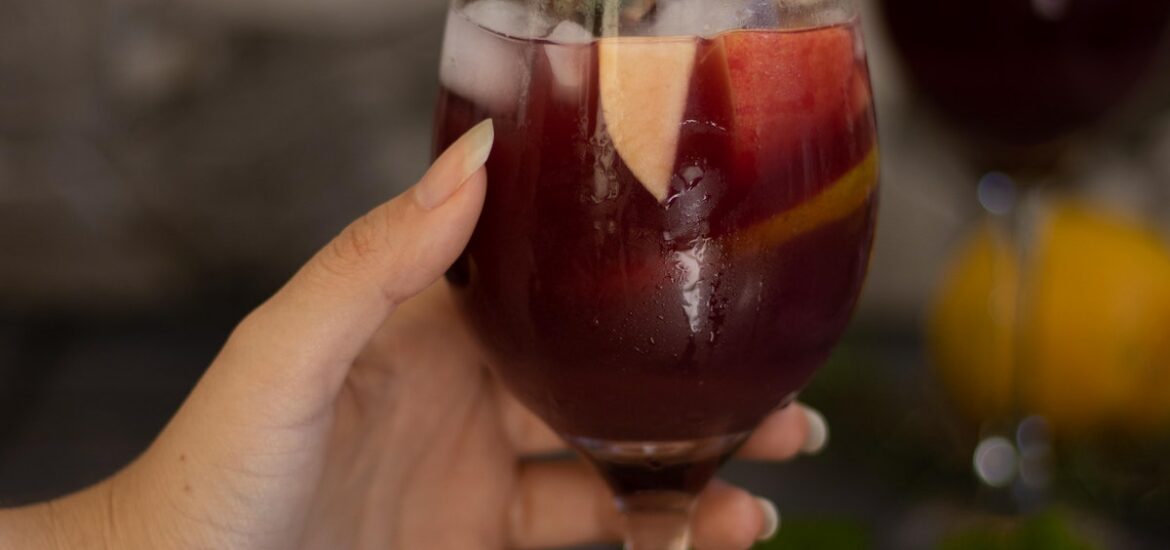 Person's hand holding a wine glass of sangria garnished with fruit and fresh herbs