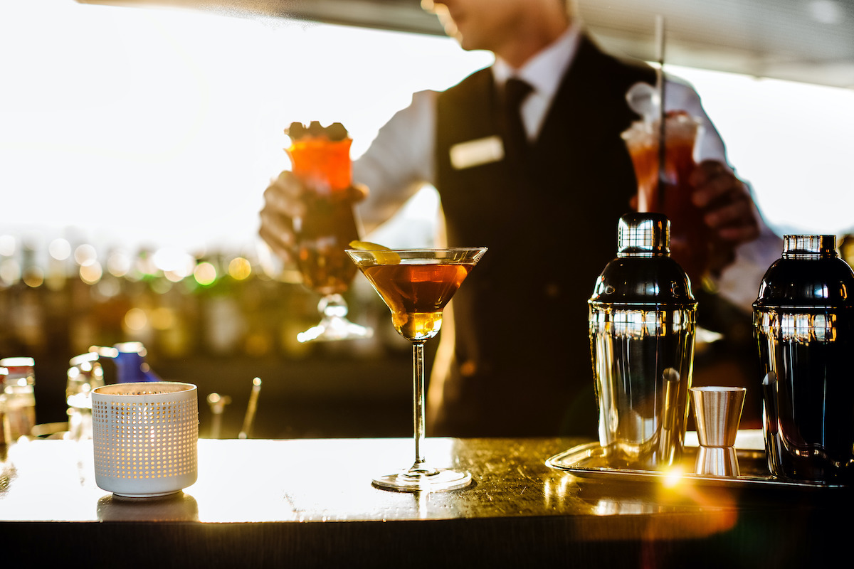 Brown cocktail in a martini glass at an outdoor terrace with a uniformed bartender in the background