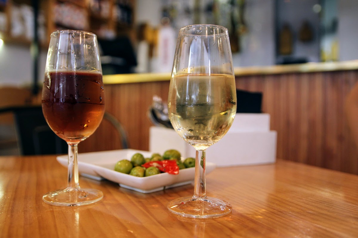 Two glasses of sherry wine (one amber-colored and one light yellow) in front of a dish of olives on a wooden tabletop.