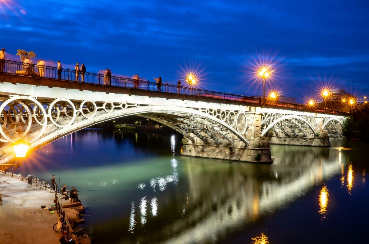 The Triana bridge illuminated at night as people sit by the river in Seville. 