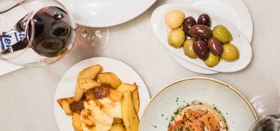 Overhead shot of various small plates of food and glasses of red wine on a white tabletop.