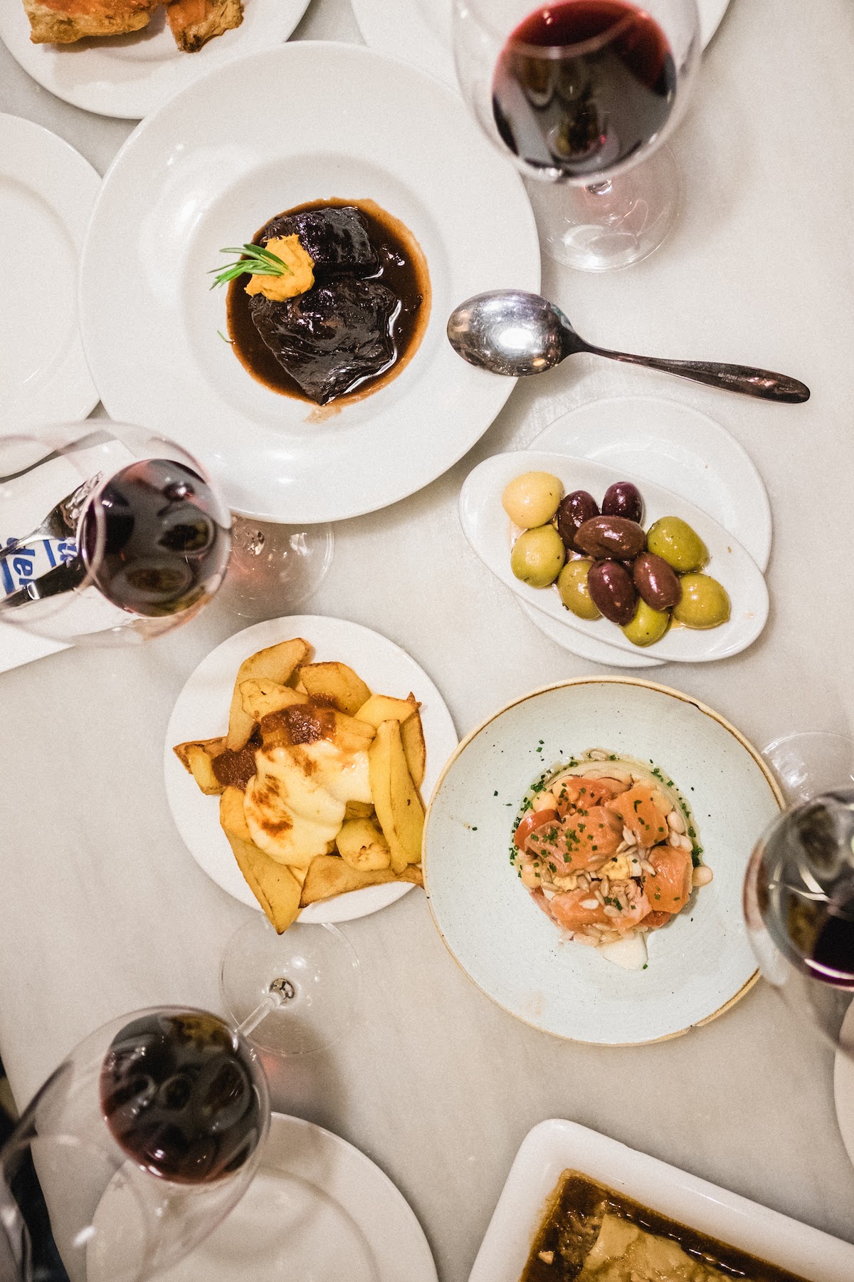 Overhead shot of various small plates of food and glasses of red wine on a white tabletop.