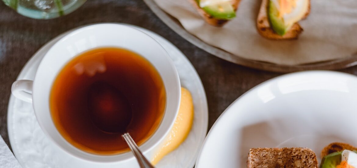 Overhead shot of a cup of tea on a saucer with a lemon slice and a plate and tiered tray with small sandwiches.