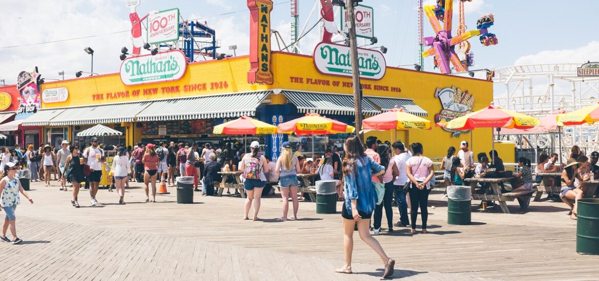 Large yellow hotdog restaurant on a boardwalk by the water in summer with many tourists and birds