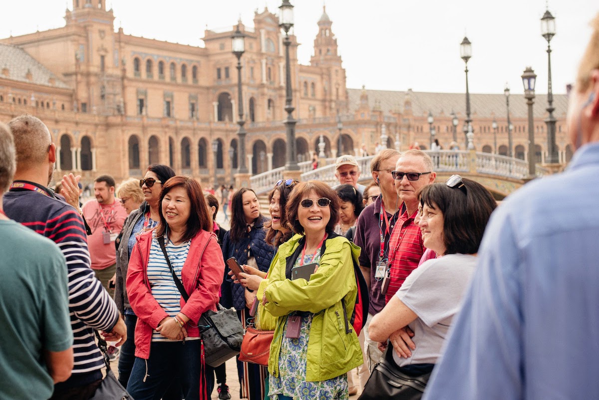 A group of tourists talking and laughing in a large plaza surrounded by brown stone buildings.