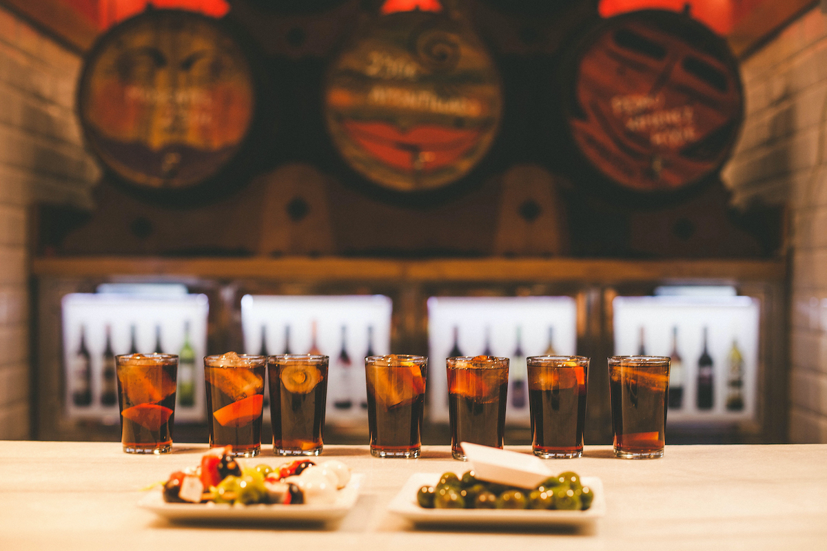 Several glasses of red vermouth and two dishes of olives and pickled skewers on a marble bar top.