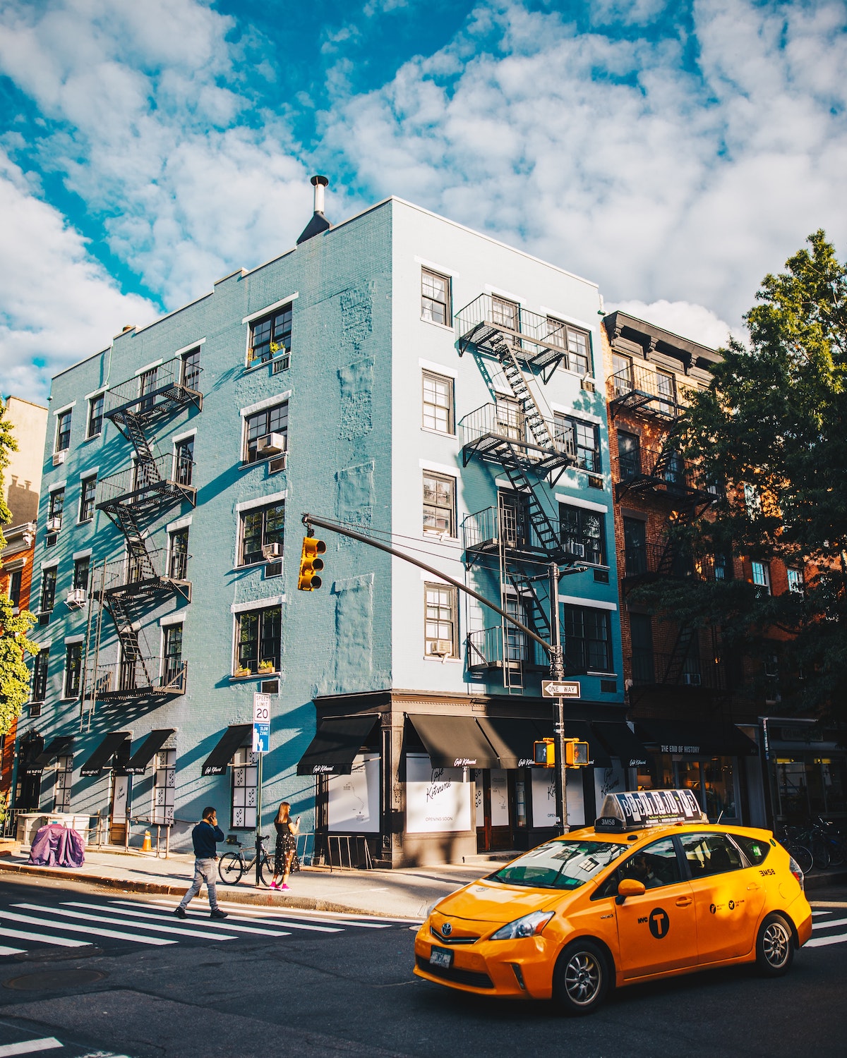 Pale blue building on a street corner with a yellow cab driving by outside.