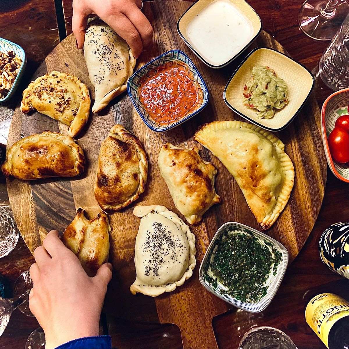 A circular tray with sauces and an array of empanadas as hands reach for them. 